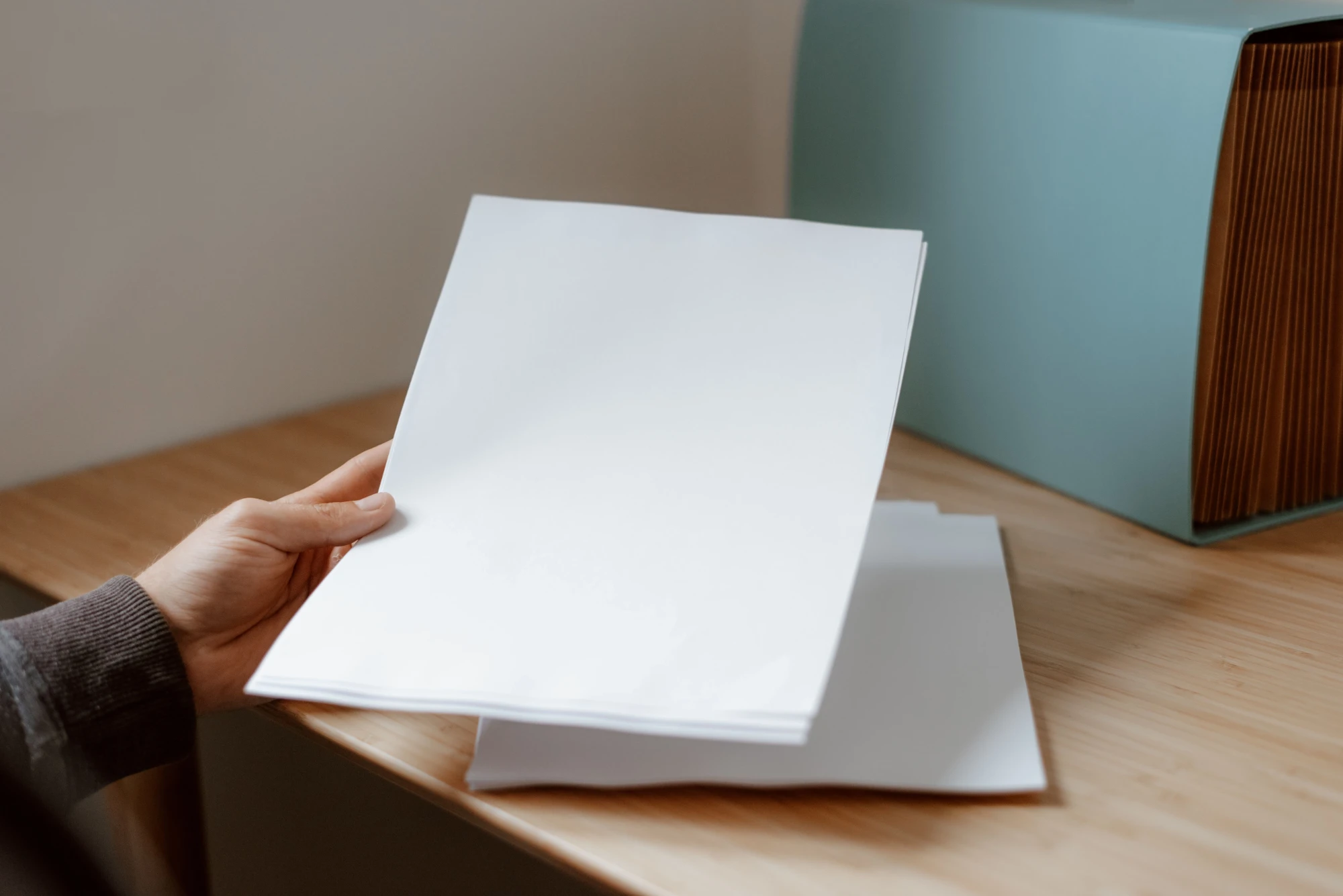 A hand lifting a stack of plain white sheets from a wooden desk